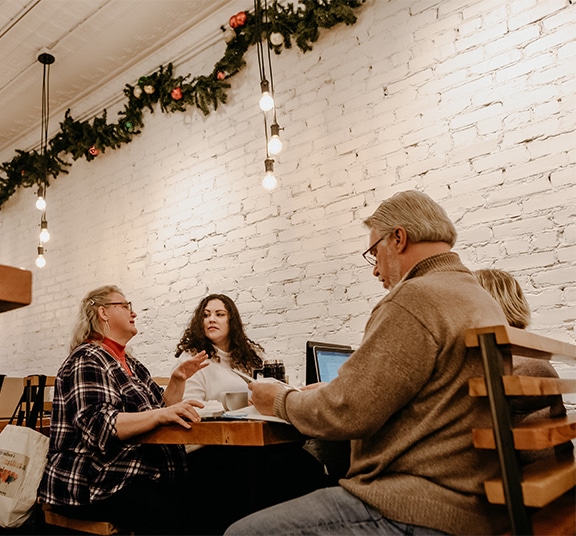 Four adult students of gender diversity talking and sitting at a coffee shop table with open books and laptops
