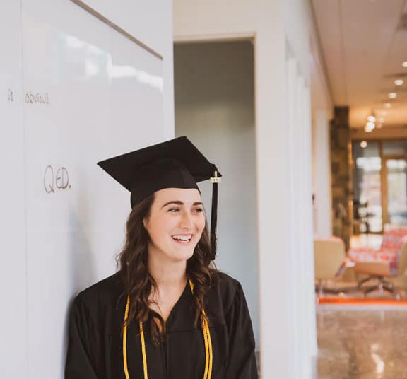 Female college graduate in cap and gown while leaning on a whiteboard in an academic building