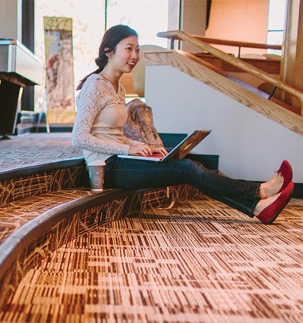Female student sitting on the steps in the Corum Student Union at Cornerstone University and typing on a laptop