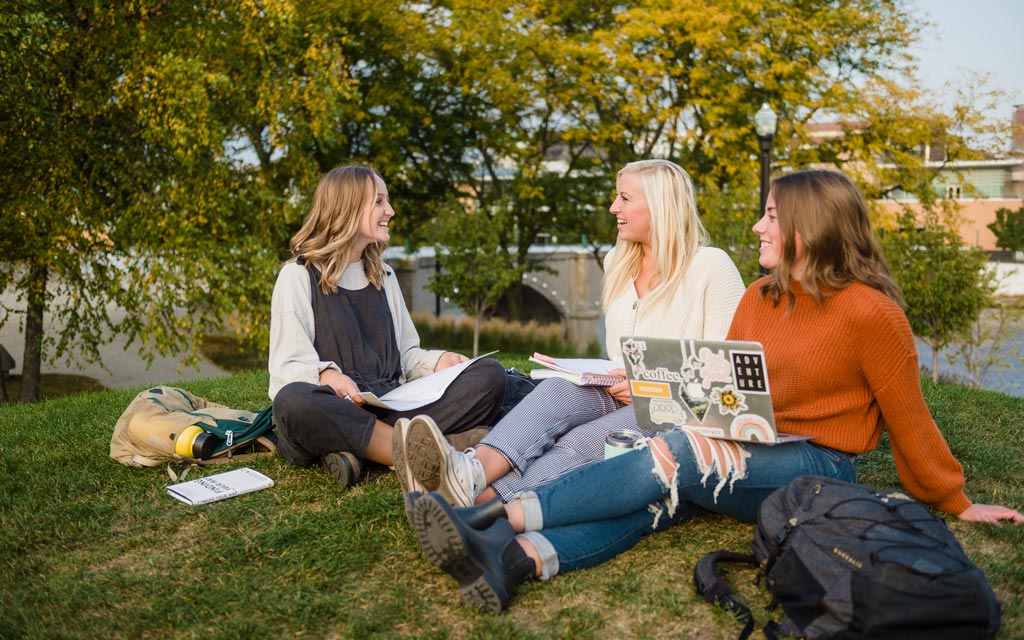 Three female college students studying on grassy knoll in Grand Rapids with a bridge in the background