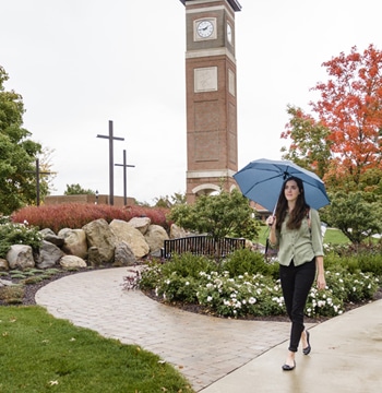 Female college student walking with an umbrella away from the bell tower on the campus of. Cornerstone University