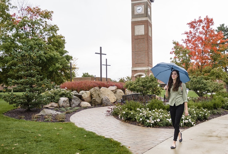 Female college student walking with an umbrella away from the bell tower on the campus of. Cornerstone University